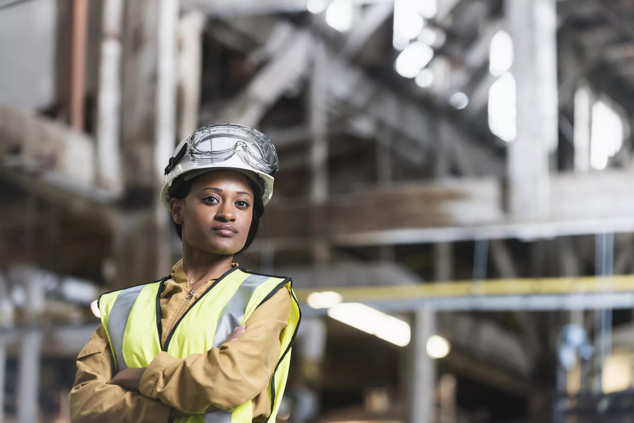 Portrait of a confident, young African American woman working in a manufacturing plant. She is standing with arms folded, looking at the camera, in a storage facility or warehouse. She is wearing a white hardhat and yellow safety vest.